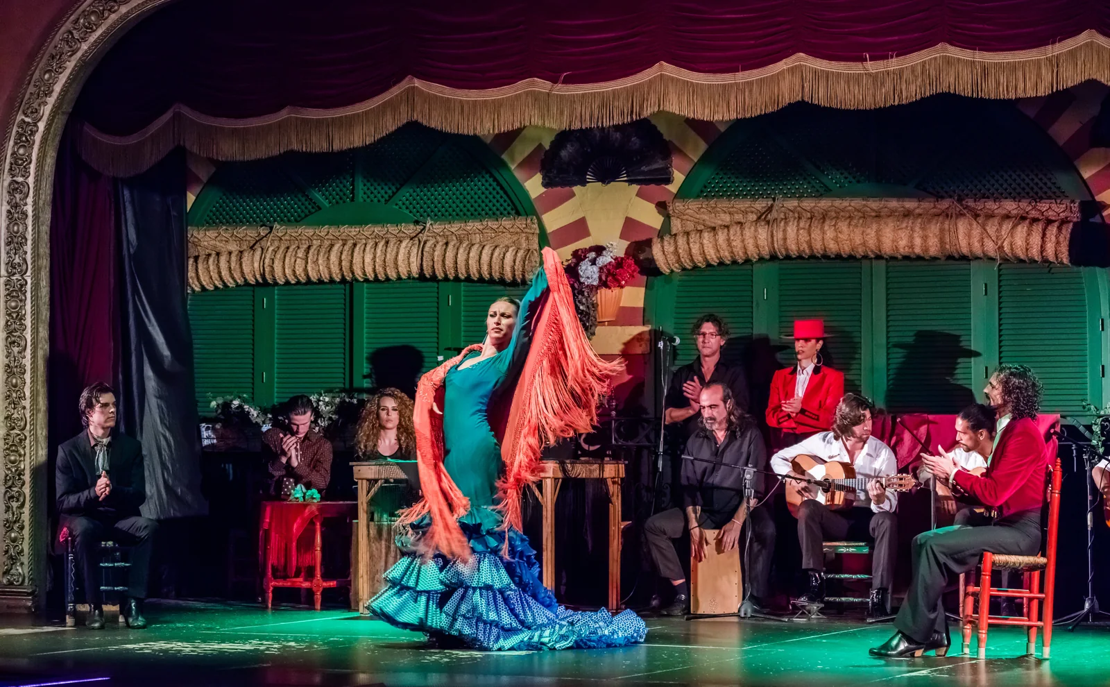 A flamenco dancer in a turquoise dress and an orange fringed shawl turns on a stage lit in green, with seated guitarists, singers and palmeros behind her under a red velvet curtain and horseshoe arches